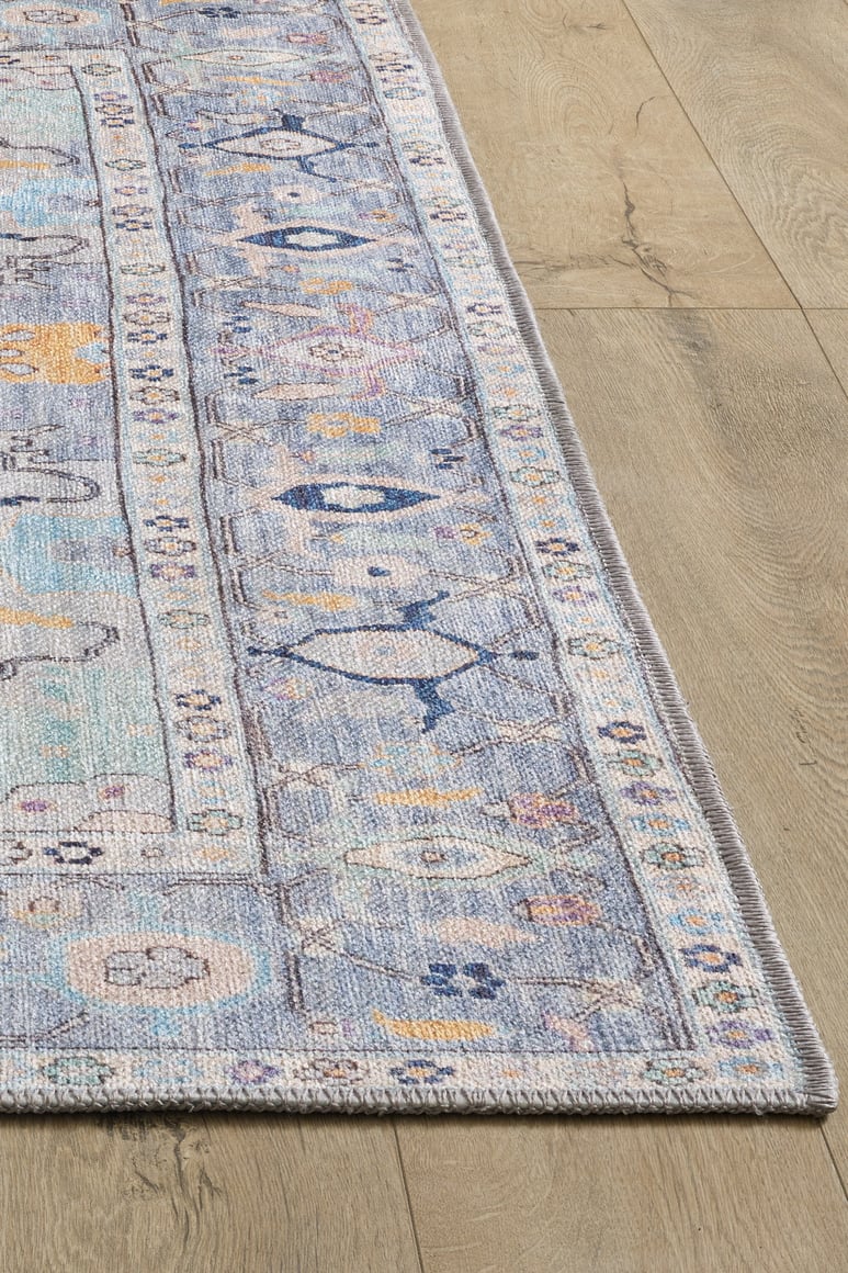 A close-up of the Seraphix Pastel Bordered Washable Rug - LCC3011 shows its rectangular, pastel blue design with a decorative border on light wood flooring—perfect for high-traffic areas with soft beige and pink intricate details.