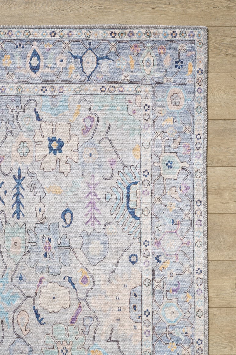 The Seraphix Pastel Bordered Washable Rug - LCC3011 features soft blue, lavender, and cream tones with floral and geometric designs plus a decorative border, shown on a light wood floor—ideal for high-traffic areas.