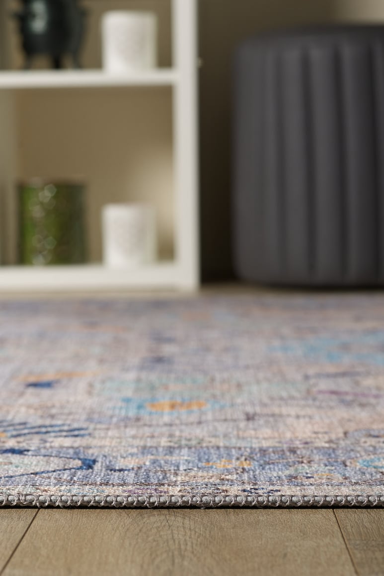 A close-up of the Seraphix Pastel Bordered Washable Rug - LCC3011 showcases its textured, multicolored design on a wooden floor, with a white shelf and dark gray ottoman in the background—ideal for high-traffic areas.