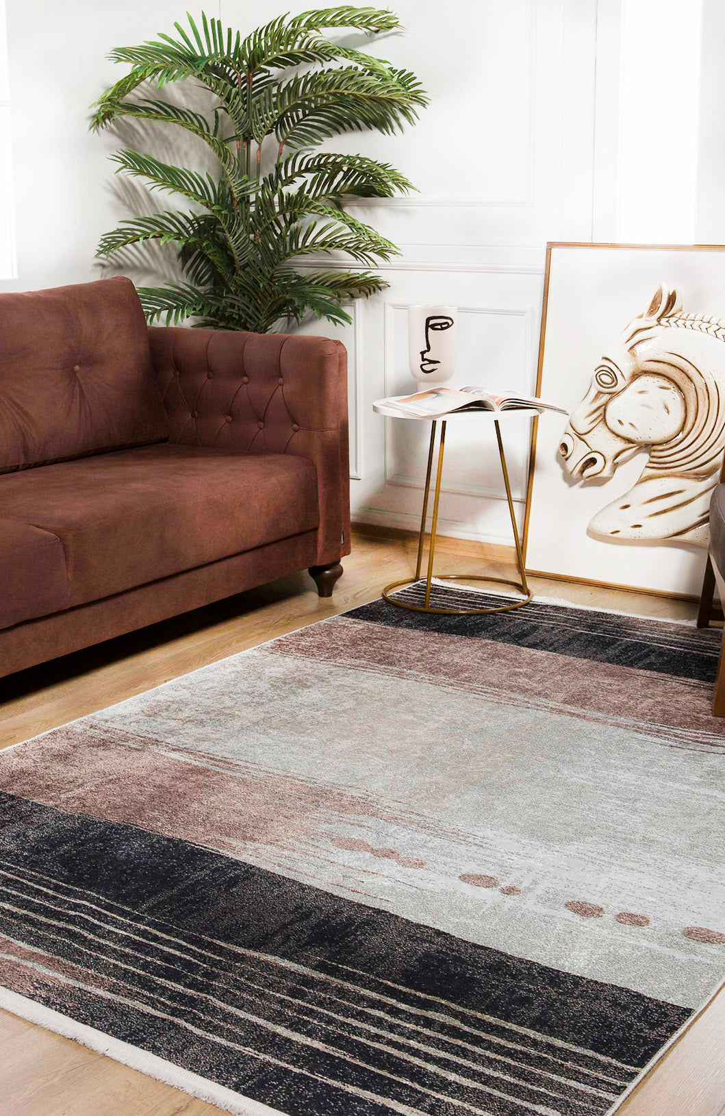 A modern living room with a brown sofa, gold and white side table, potted plant, large abstract horse artwork on the floor, and the Serene Swirls - Washable Rug - JR952 (Custom Size) in neutral tones—ideal for high-traffic spaces.