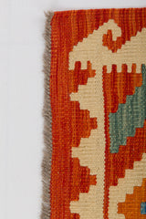 A close-up of the Silk Road - Handwoven Kilim (Outlet) US shows its geometric patterns in orange, cream, and green, a textured flatweave surface, and fringed left edge, all set against a plain white background.