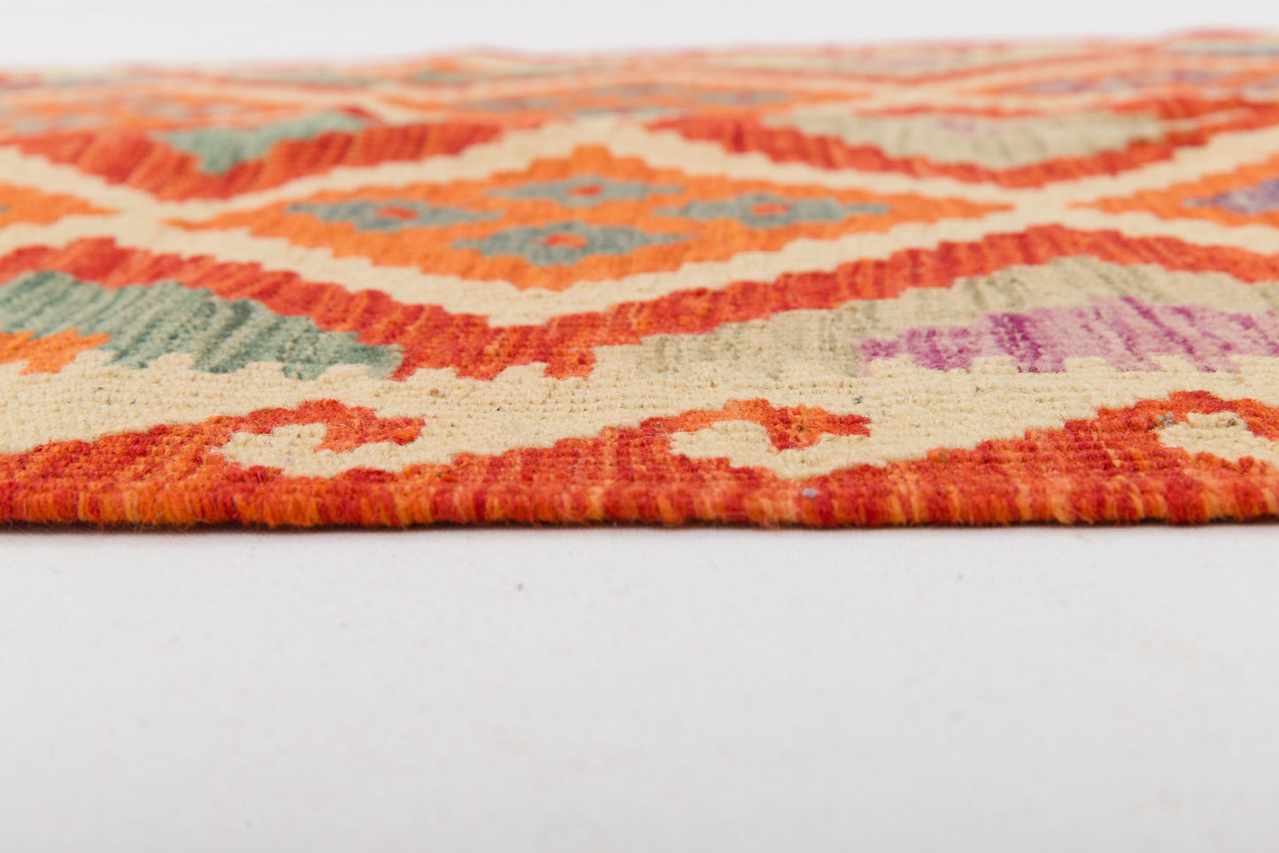 A close-up side view of the Silk Road - Handwoven Kilim (Outlet) US, showcasing geometric diamond and swirl patterns in orange, cream, green, and purple on a white surface.