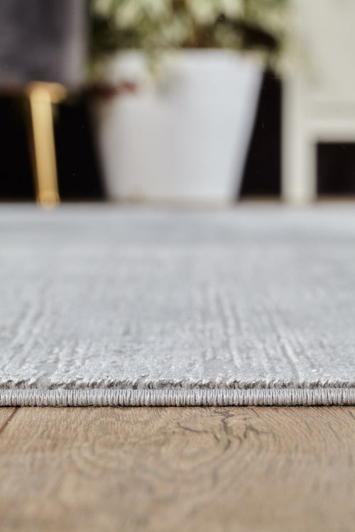 Load image 1 in gallery view. Close-up of the edge of the Silver Lining Grey Rug - M352I on a wooden floor, with a white plant pot and blurred background furniture visible in the distance.