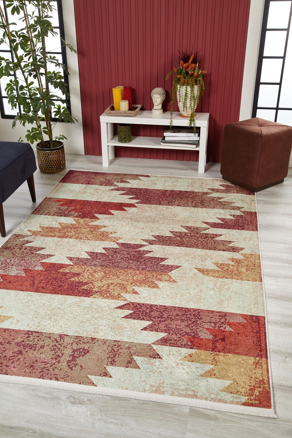 A living room featuring the Simplistic Artistry - Washable Rug - JR1647 (Custom Size) in geometric red, orange, and cream tones, a white console table with decor, potted plant, brown ottoman, and a red accent wall. Natural light fills the space.