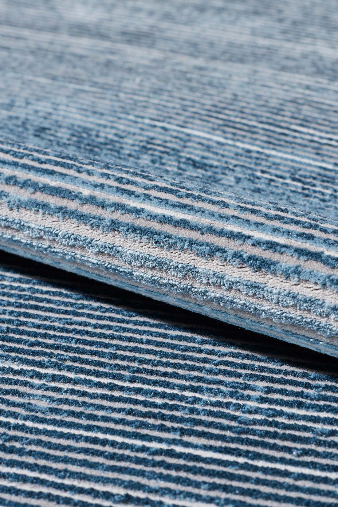Close-up of the Slate to Midnight Navy Rug - M497M, a machine-made piece with textured fabric and horizontal blue and white stripes, shown with a rolled edge atop a matching striped surface.
