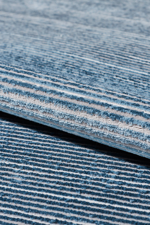 Load image 1 in gallery view. Close-up of the Slate to Midnight Navy Rug - M497M, a machine-made piece with textured fabric and horizontal blue and white stripes, shown with a rolled edge atop a matching striped surface.