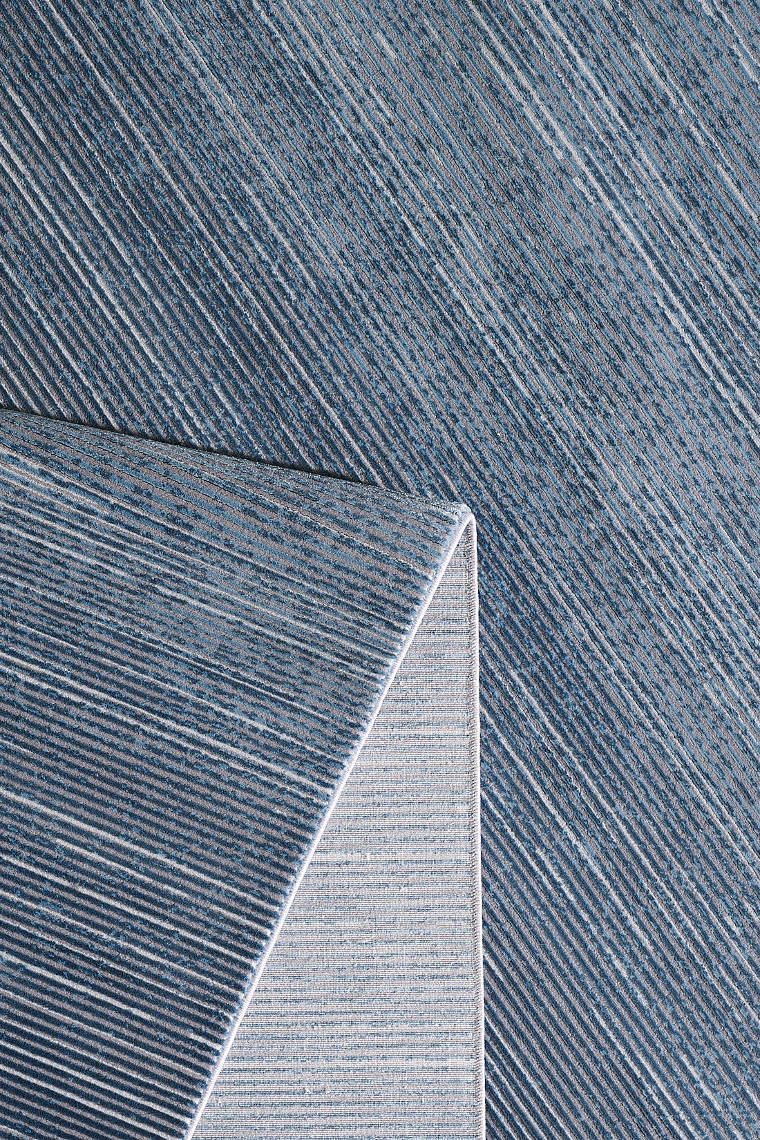 A close-up of the Slate to Midnight Navy Rug - M497M shows blue gradients with diagonal white stripes; one corner is folded to reveal a lighter striped pattern on the underside.