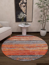 A modern living room features the Sleek Brushed - Washable Rug - JR431 (Custom Size) in orange and gray, ideal for high-traffic areas, along with a small white side table holding books and flowers, a potted plant, a beige sofa, and classical sculpture wall art.