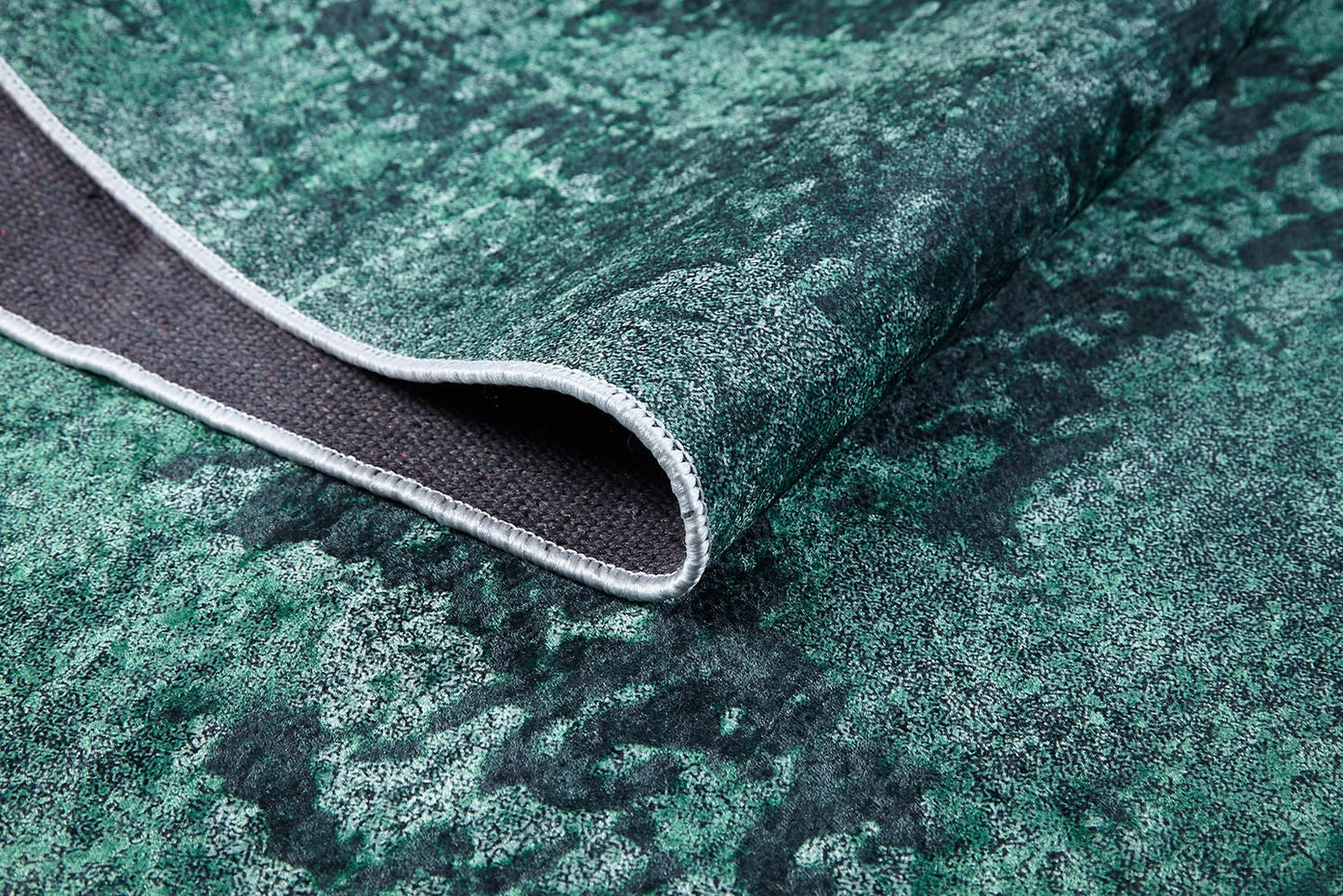 A close-up of the Smokey Mountains - Washable Rug - JR1631 (Custom Size) in green and black, with one corner bent to reveal its dark woven underside and stitched edge.