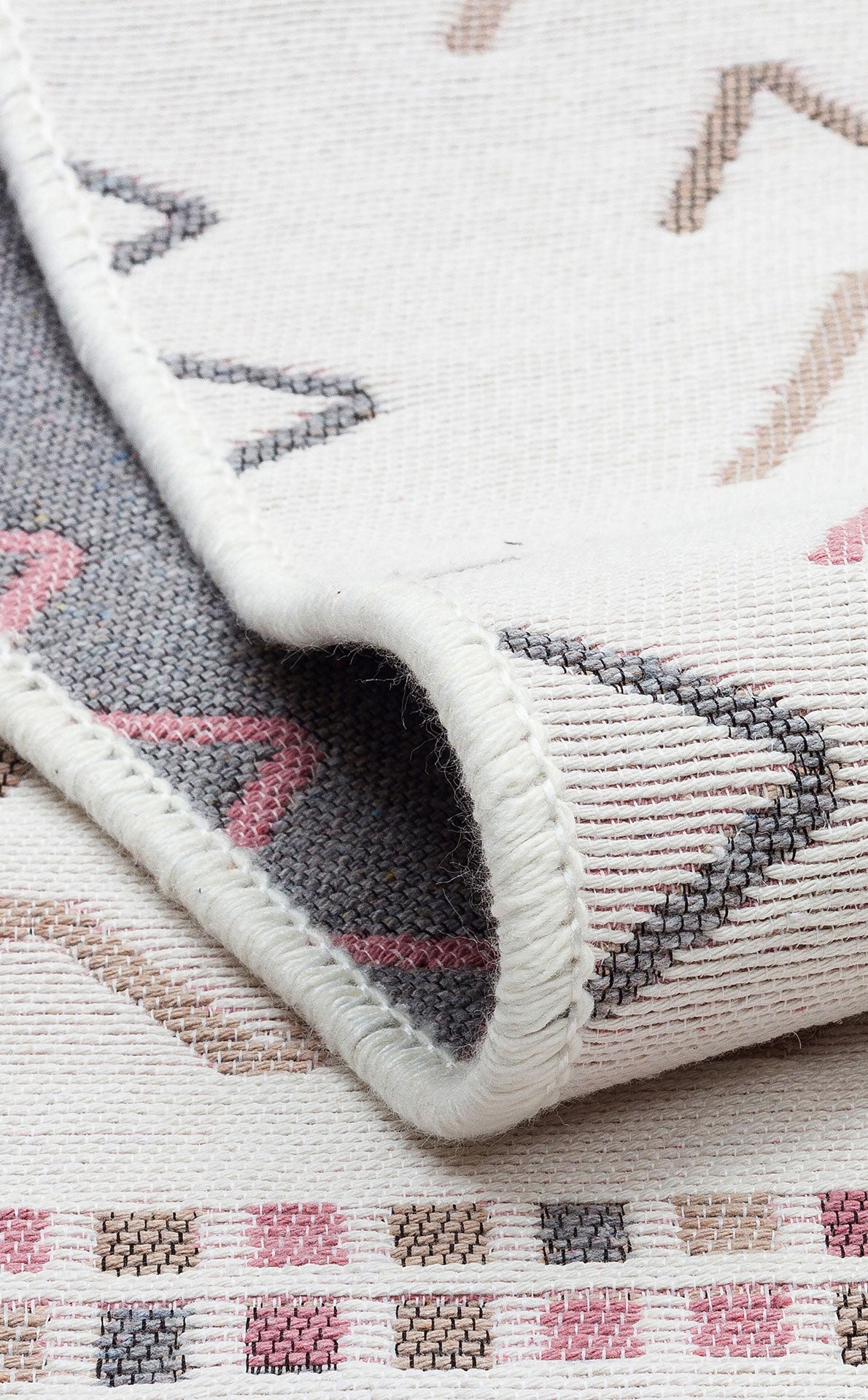 A close-up of the Soft Bloom - Washable Double-Sided Kilims - AR05 shows abstract geometric patterns in pink, grey, and beige. The partially folded rug highlights its texture and detailed stitching.