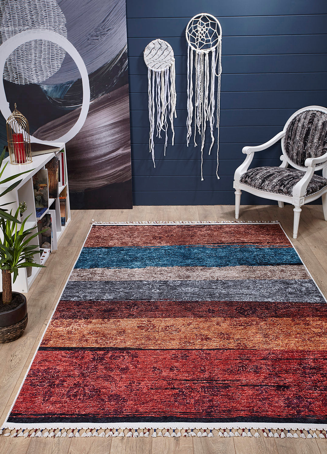 A modern living room showcases the Soft Brushed - Washable Rug - JR5056 (Custom Size), a white armchair with a patterned cushion, dreamcatchers on a dark blue wall, a white bookshelf, a potted plant, and wood floors—ideal for busy spaces.
