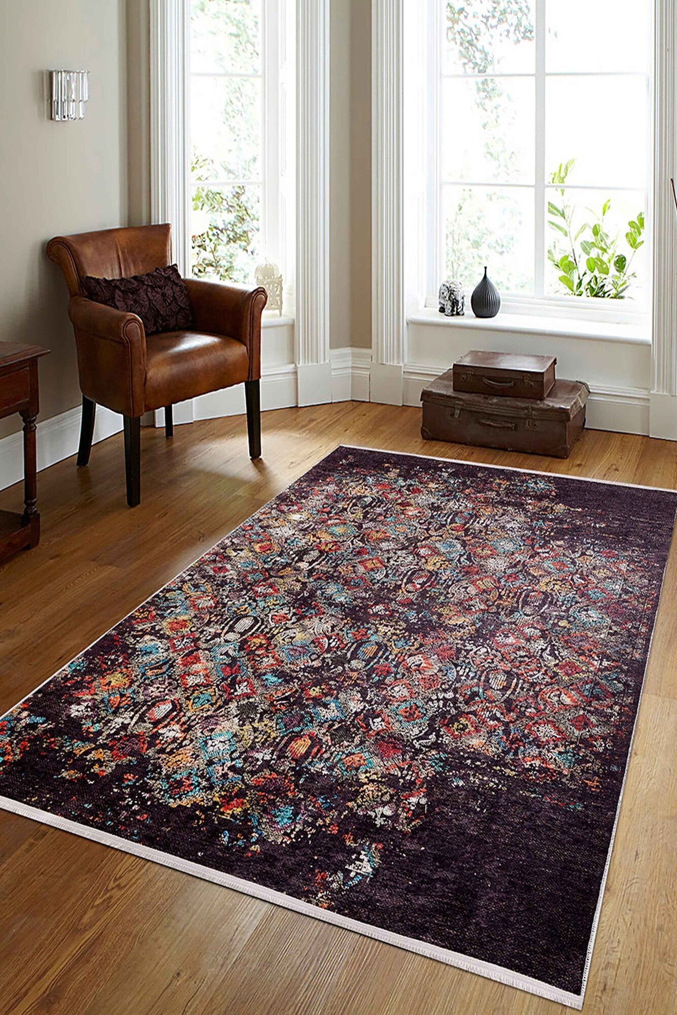 A cozy living room with wooden floors, the Soft Haze - Washable Rug - JR1094 (Custom Size) in red, blue, and purple—ideal for high-traffic spaces—a brown leather armchair, small side table, stacked suitcases, and sunlit windows.