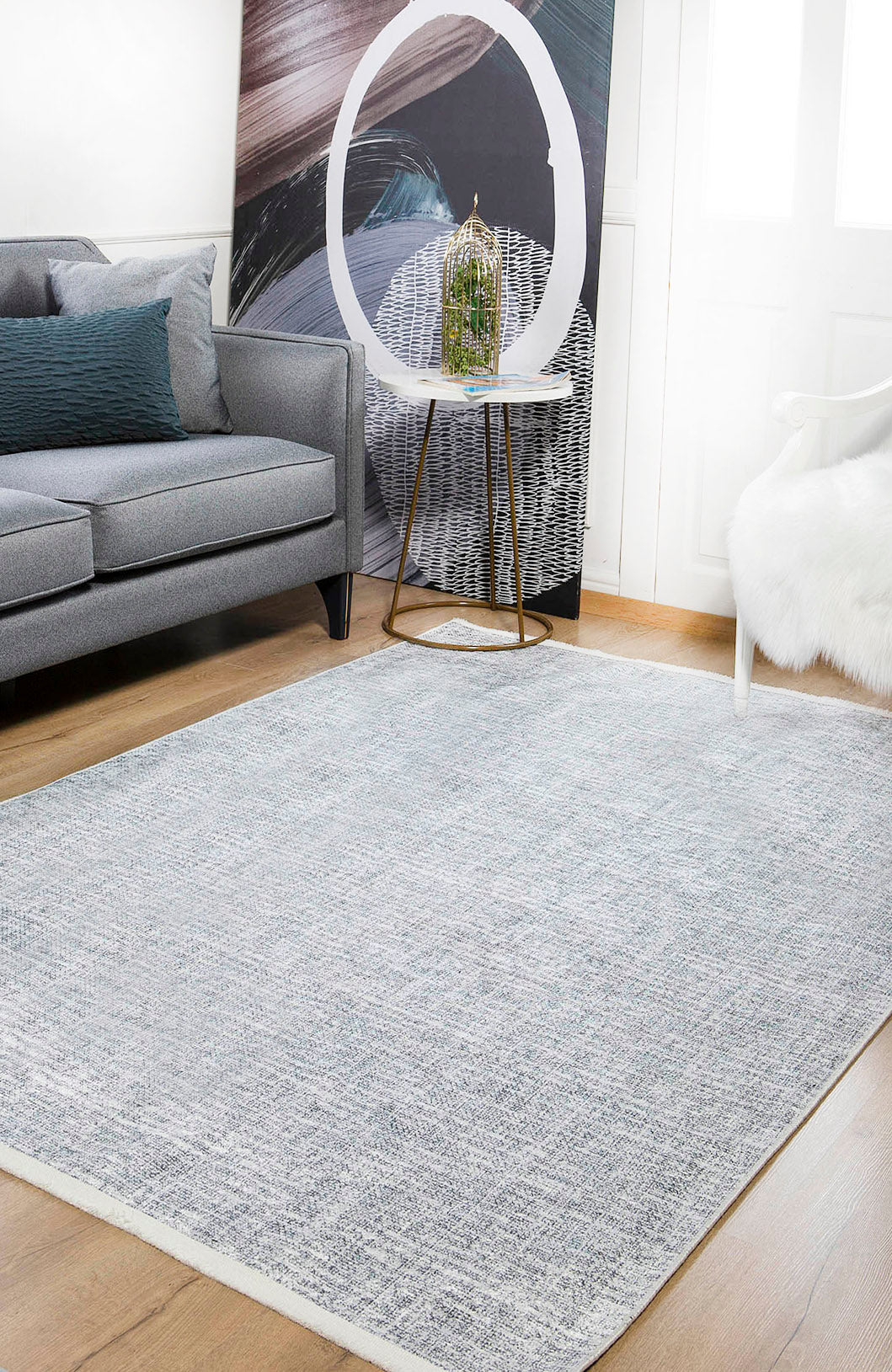 A modern living room with a gray sofa, white furry chair, and the Softly Chic - Washable Rug - JR747 (Custom Size) in gray-anthracite on wooden floors, plus a small round side table with decor and abstract art against the wall.