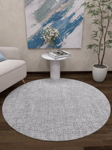 A modern living room featuring the Softly Chic - Washable Rug - JR747 (Custom Size) in gray-anthracite, a white side table with books and flowers, a potted plant, abstract wall art, and part of a light sofa on a dark wood floor.