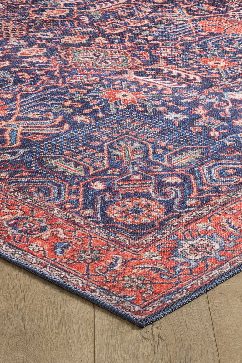 The Solstice Red Navy Washable Rug (LCC3026) with vivid geometric and floral patterns in red, navy, orange, and purple is shown close-up on a wooden floor, its allergy-friendly design featuring a pointed corner in the foreground.