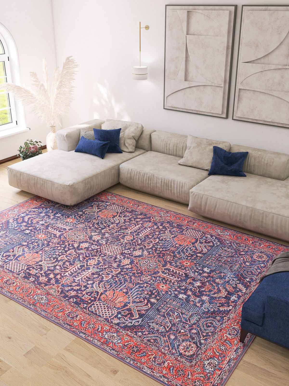 A modern living room features a large beige sectional, blue accent pillows, abstract wall art, tall white pampas grass, and the Solstice Red Navy Washable Rug - LCC3026 (Custom Size), perfect for high-traffic areas on light wood floors.