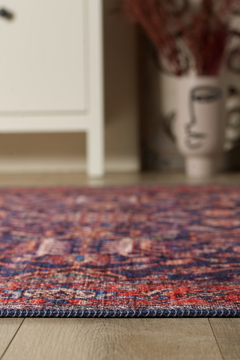 A close-up of the Solstice Red Navy Washable Rug - LCC3026 (Custom Size) featuring vibrant red and blue tones on a wooden floor, ideal for high-traffic areas, with a cream cabinet and decorative vase nearby.