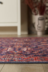 A close-up of the Solstice Red Navy Washable Rug - LCC3026 (Custom Size) featuring vibrant red and blue tones on a wooden floor, ideal for high-traffic areas, with a cream cabinet and decorative vase nearby.