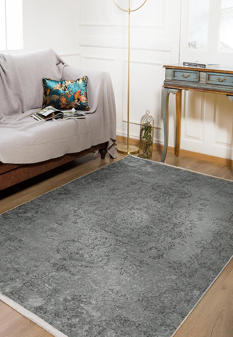 A cozy living room with a brown sofa, a light gray throw, a colorful cushion, the Sophisticated Design - Washable Rug - JR173 (Custom Size) in gray-anthracite for high-traffic areas, a blue vintage console table, floor lamp, white paneled walls, and wooden flooring.