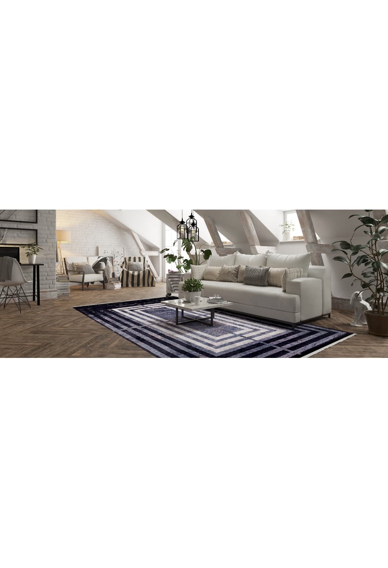 A modern loft living room features a white sofa, neutral cushions, a glass coffee table, and the Sophisticated and Chic - Washable Rug - JR1409. Potted plants, large windows, and exposed wooden beams create an airy, charming space.