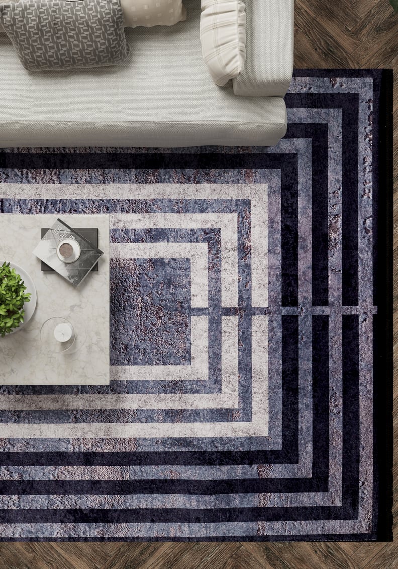 A modern living room features the Sophisticated and Chic - Washable Rug - JR1409 (Custom Size) with concentric rectangles in blue and gray, a marble coffee table with decor, and a light gray sofa accented by patterned pillows.