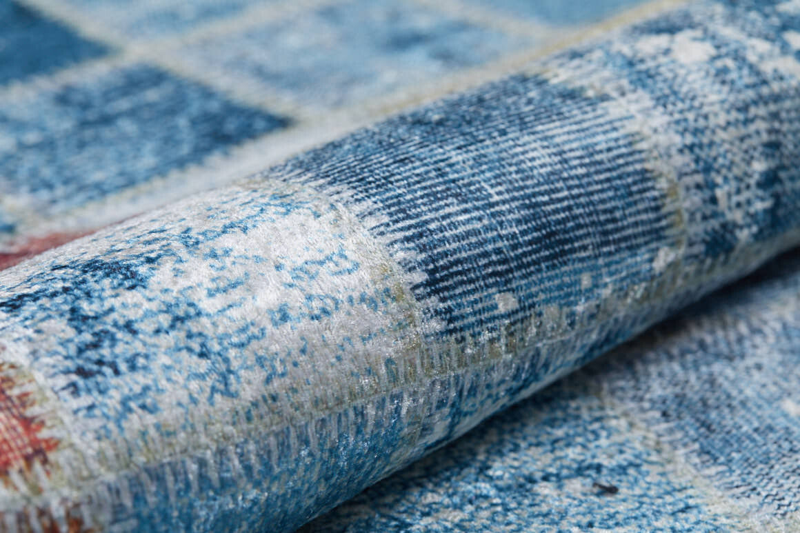 A close-up of the Square Serenity - Washable Rug - JR1255 (Custom Size) shows its blue and white patchwork design, textured surface, and intricate details. This washable rug is partially rolled and ideal for high-traffic areas.