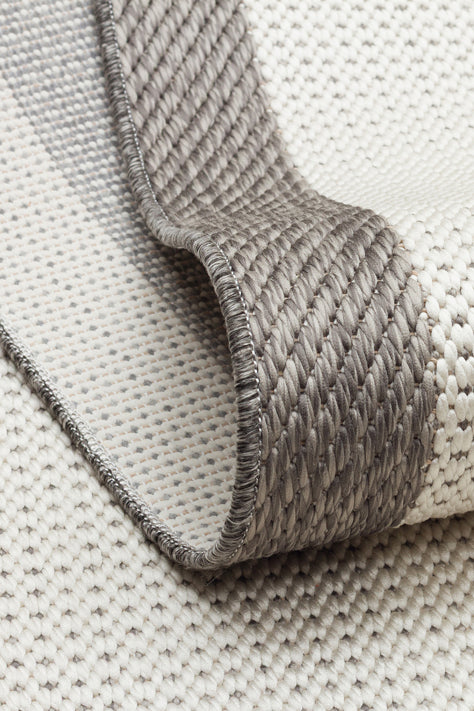 A close-up of the Stone Layer - Kilim - MNO04 flatwoven polyester rug in beige and gray, highlighting its textured surface, folded edge, detailed stitching, and the underside.