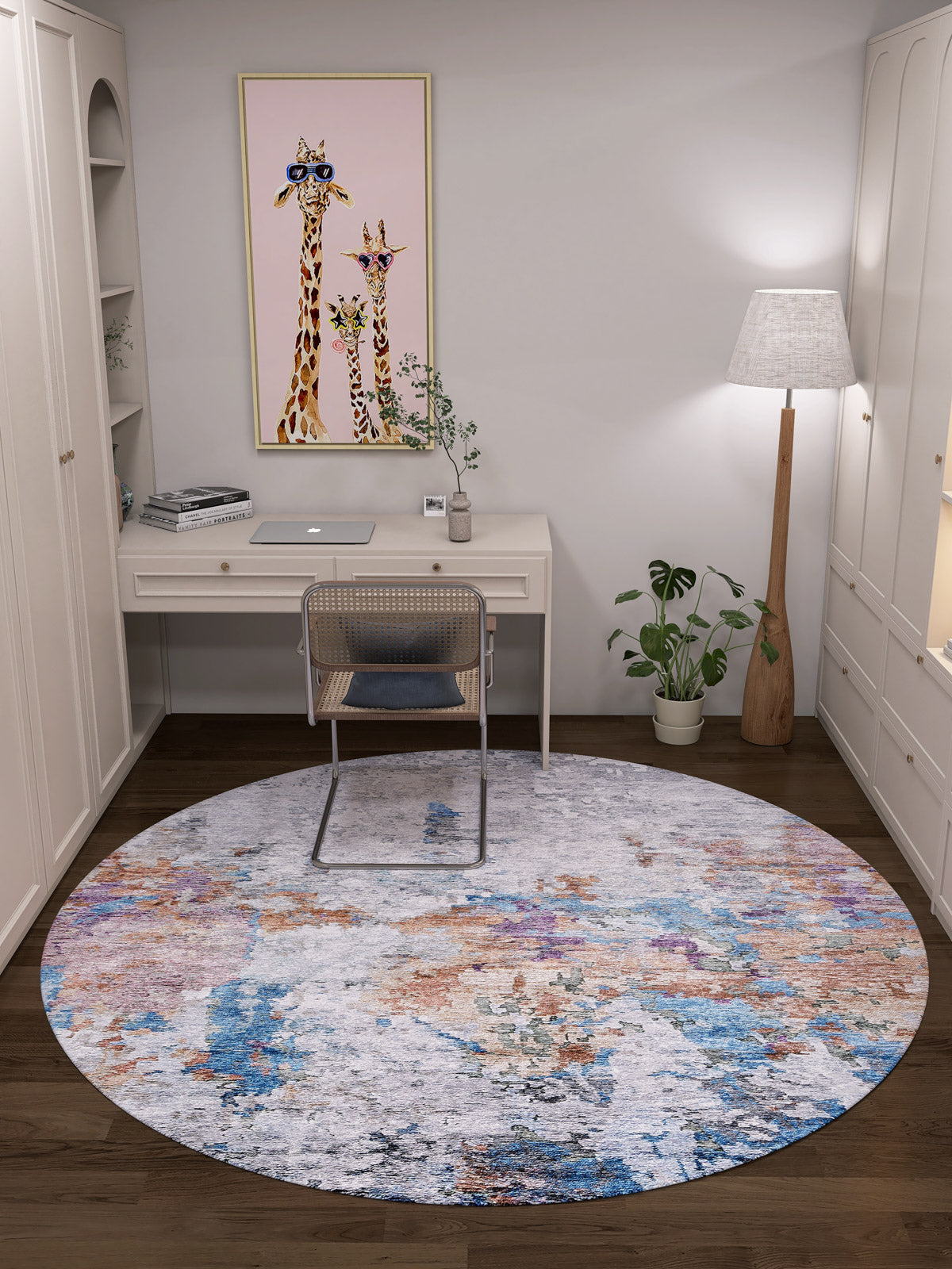 A modern home office features a white desk, rattan chair, laptop, and the Striking Statements - Washable Rug - JR786. A floor lamp, potted plant, giraffe artwork, white shelves, and cabinets complete the cozy, well-lit space.