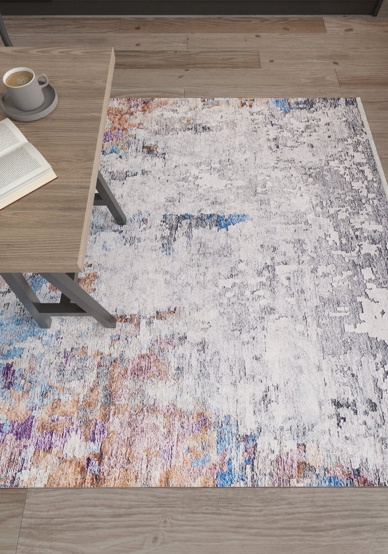 The Striking Statements - Washable Rug - JR786 (Custom Size), featuring modern abstract multicolor tones, lies on a wooden floor beside a light wood table with an open book and a cup of coffee.