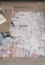 The Striking Statements - Washable Rug - JR786 (Custom Size), featuring modern abstract multicolor tones, lies on a wooden floor beside a light wood table with an open book and a cup of coffee.