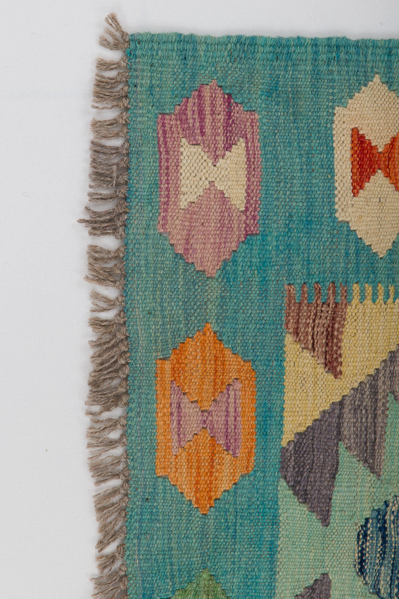 A close-up of the Striped Elegance - Handwoven Kilim, featuring a teal background, orange, purple, and red geometric patterns, and fringed edges on the left side.