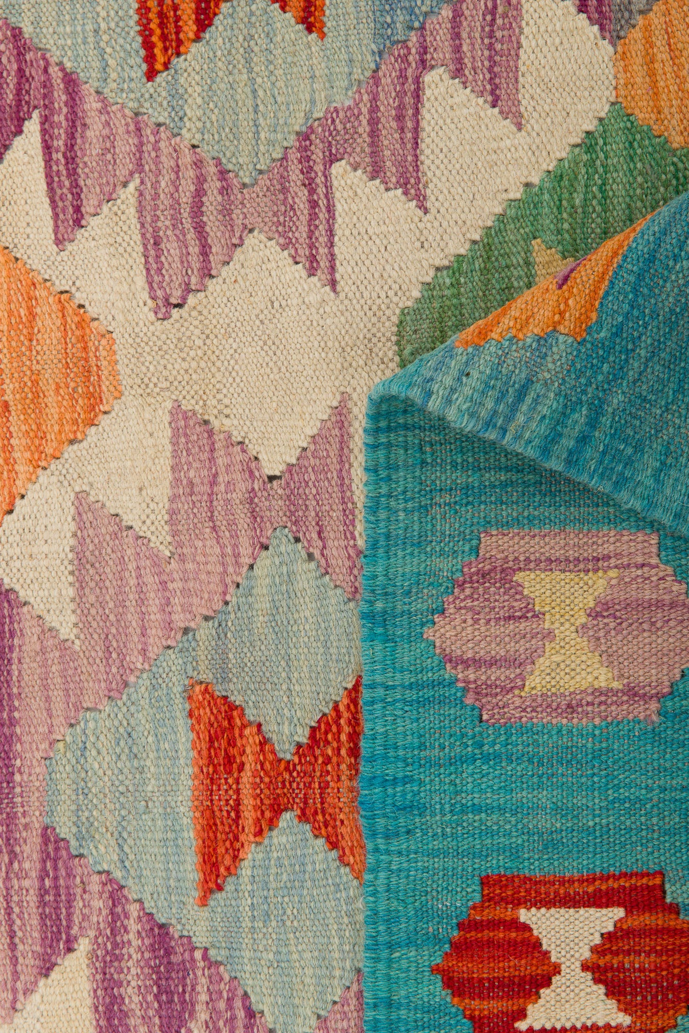 The Striped Elegance - Handwoven Kilim features vivid geometric patterns in orange, purple, red, and cream, with a folded corner showing a blue-green flatweave underside decorated with diamond shapes.