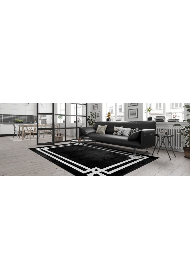 A modern living room features a black sofa, decorative pillows, and the Subtle Border - Washable Rug - JR597 (Outlet) US in black with white geometric patterns. Large windows, plants, and a dining area visible in the background brighten the space.