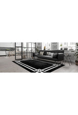 A modern living room features a black sofa, decorative pillows, and the Subtle Border - Washable Rug - JR597 (Outlet) US in black with white geometric patterns. Large windows, plants, and a dining area visible in the background brighten the space.