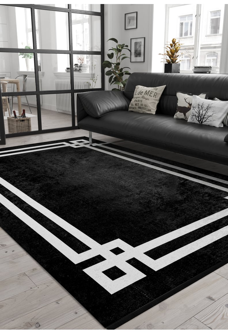 A modern living room with a black sofa, decorative pillows, potted plants, wooden floors, and large windows features the Subtle Border - Washable Rug - JR597 (Custom Size), perfect for high-traffic areas. A black-framed glass wall divides the space.