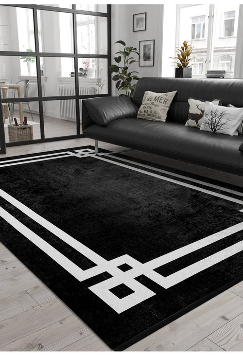Bild 1 in Galerieansicht laden. A modern living room with a black sofa, decorative pillows, potted plants, wooden floors, and large windows features the Subtle Border - Washable Rug - JR597 (Custom Size), perfect for high-traffic areas. A black-framed glass wall divides the space.