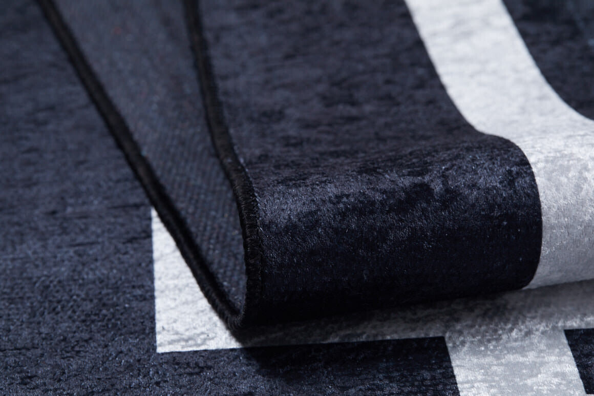Close-up of the Subtle Border - Washable Rug - JR597 (Custom Size) in black with a white geometric pattern; one corner is folded to show its thick, textured build, making it durable and ideal for high-traffic areas.