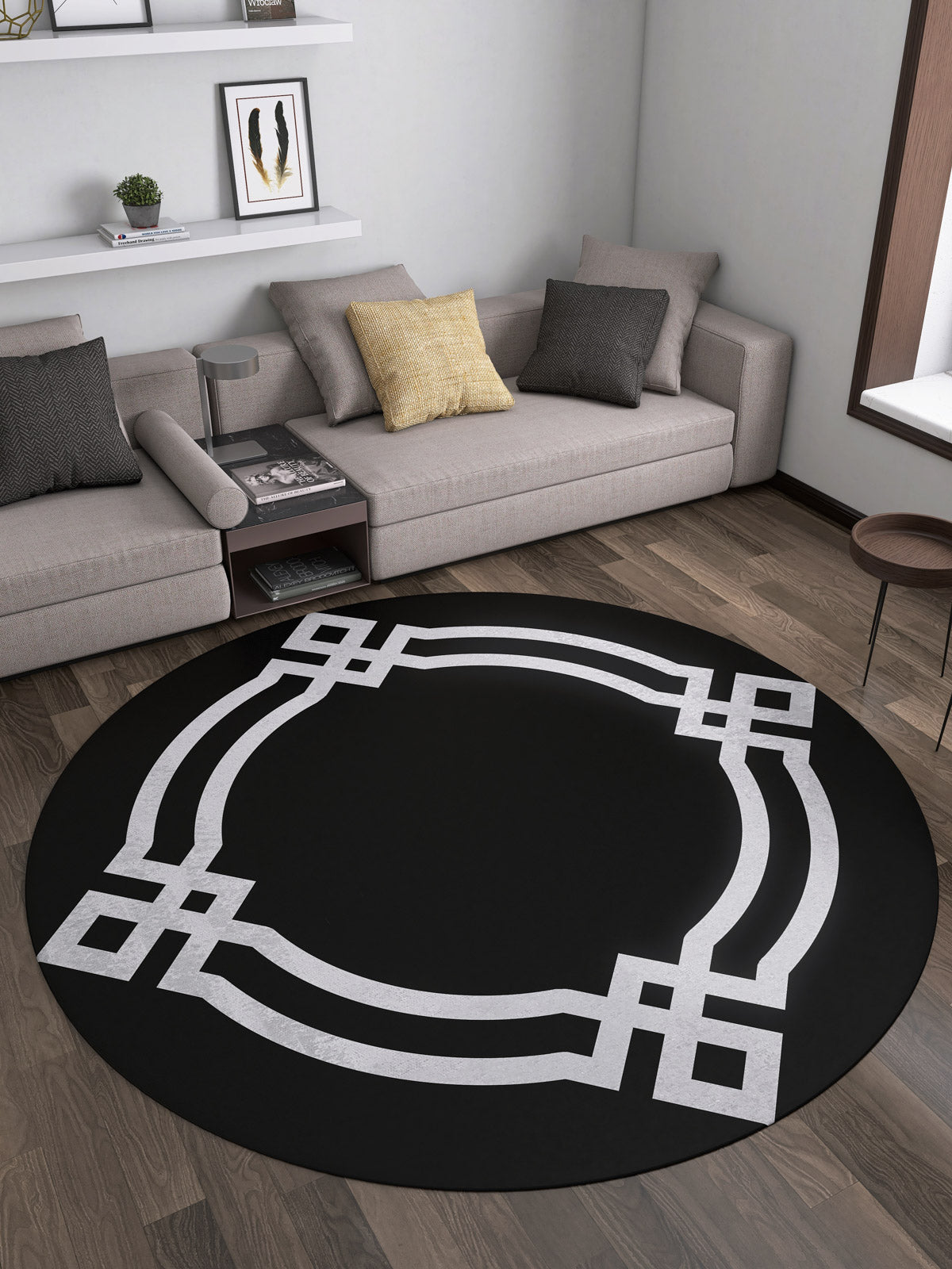 A modern living room with the Subtle Border - Washable Rug - JR597 (Custom Size), a round black rug with a geometric white pattern, ideal for high-traffic areas, paired with a beige sectional, cushions, wall shelves, and framed art.