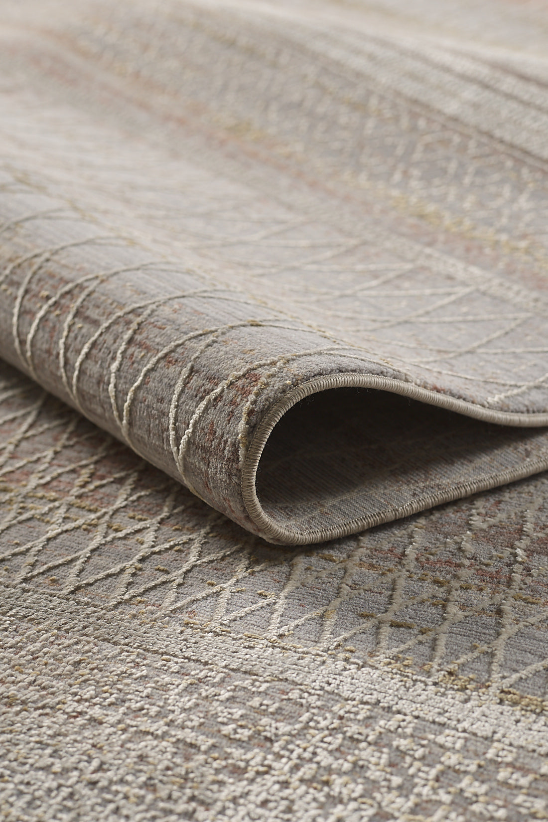 A close-up of the Sunkissed Boho Trellis Rug - M627M, rolled up to reveal its textured surface with subtle diamond and linear patterns in muted beige, gray, and taupe tones.
