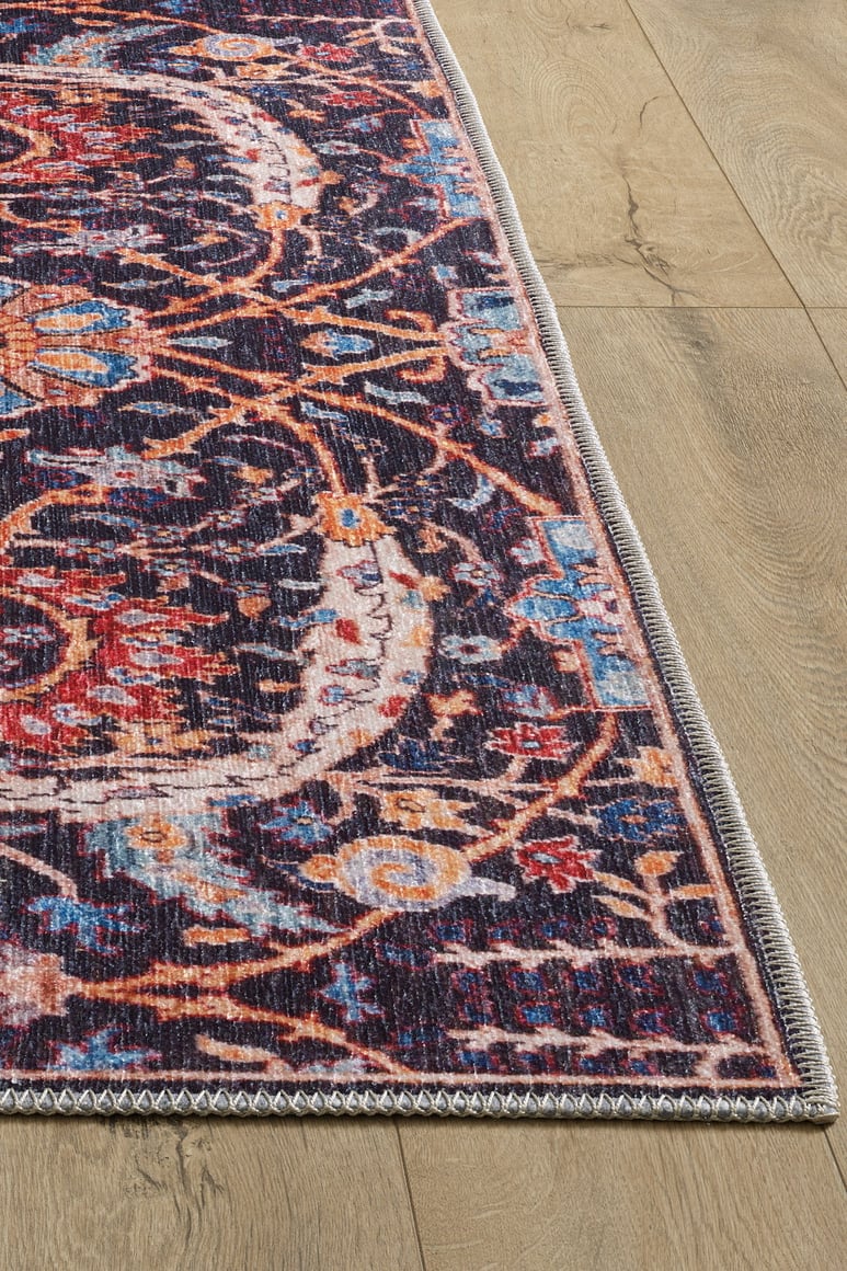 A close-up of the Sylvanor Colorful Washable Rug - LCC3017, showcasing its intricate red, blue, orange, and beige patterns on a light wood floor. This washable rug features detailed stitched edges and a vintage, slightly distressed look.