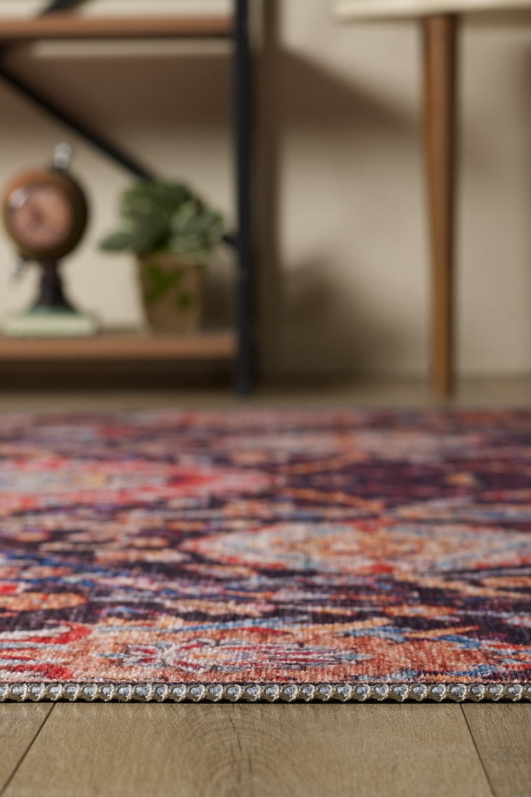 A close-up of the Sylvanor Colorful Washable Rug - LCC3017 on a wooden floor, ideal for high-traffic areas, with a potted plant, decorative clock, and wooden furniture legs in the softly blurred background.