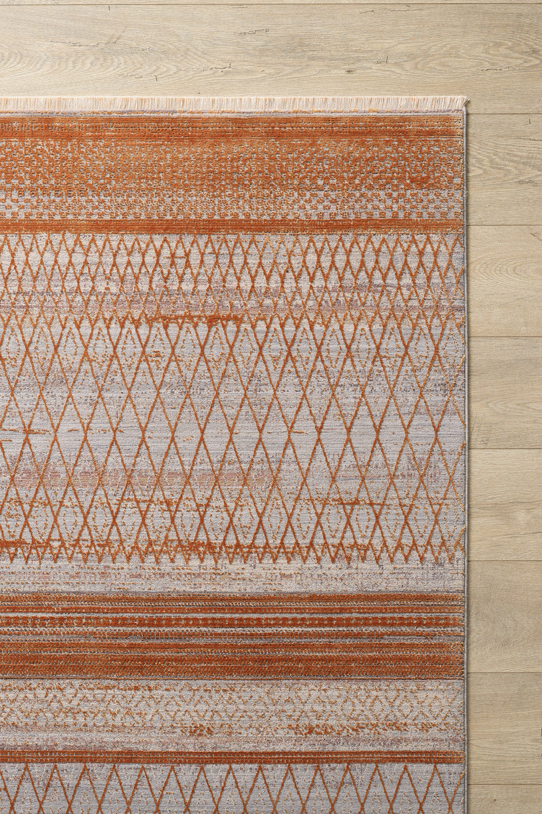 The Tangerine Dream Trellis Rug - HRD008 is a modern diamond pattern rug with orange and beige geometric designs, horizontal lines, and fringed edges on one side, placed on a light wood floor. This rug is machine-made for added durability.
