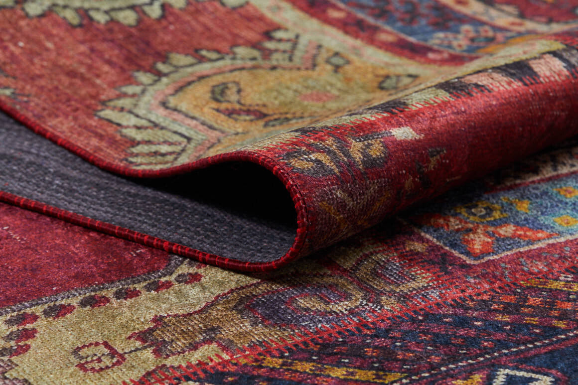 A close-up of the Tapestry Tango - Washable Rug - JR313 (Custom Size), featuring vibrant red, gold, and blue patterns with a folded corner—ideal for high-traffic spaces and shown against a textured background.