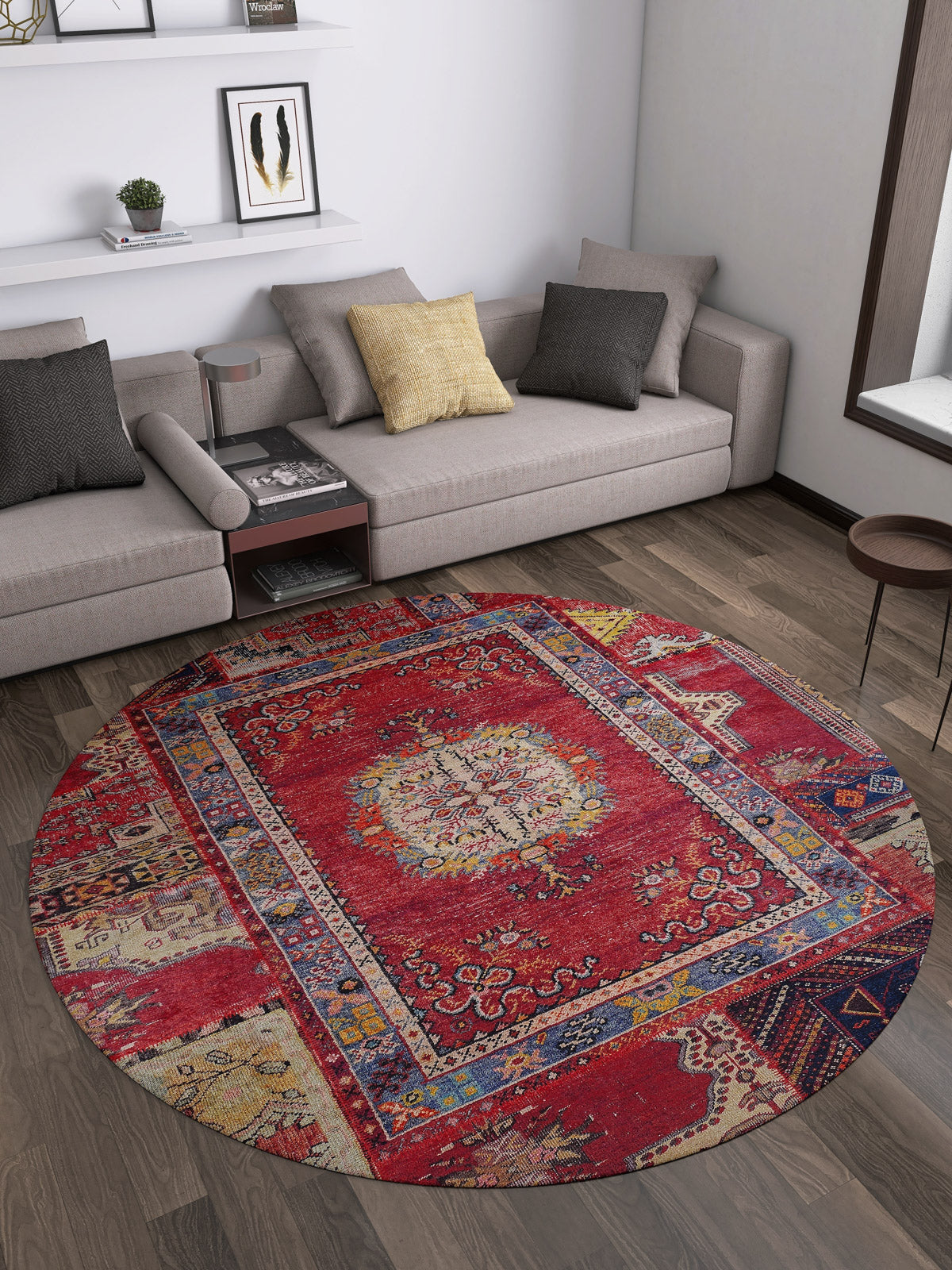 A modern living room with a gray L-shaped sofa, neutral cushions, the Tapestry Tango Washable Rug - JR313 (Custom Size) in red on dark wood floors—perfect for high-traffic areas—a small round table, wall art above a low white shelf, and a large window letting in light.