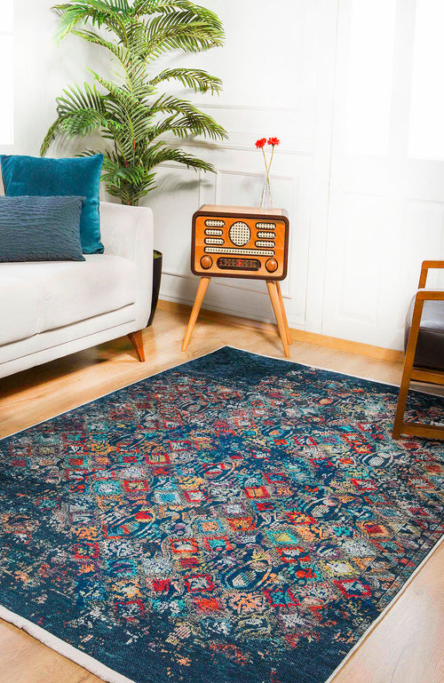 Load image 1 in gallery view. A cozy living room with the Tarnished Elegance - Washable Rug - JR1095 (Custom Size) in blue for high-traffic areas, a white sofa with blue pillows, wooden armchair, vintage radio with red flowers, potted plant, and sunlit white walls.