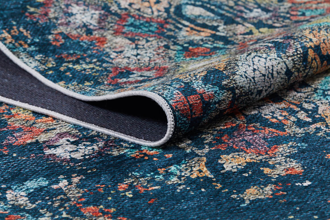 A close-up of the Tarnished Elegance - Washable Rug - JR1095 (Custom Size) in vibrant blue with teal, red, yellow, and white patterns; one corner is folded to reveal its detailed texture and dark underside—ideal for high-traffic areas.