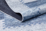 A close-up of the Tartan Twist - Washable Rug - JR31 (Custom Size), showing its blue flat weave with stitched X pattern and gray accents. The folded corner reveals the textured, washable backing.