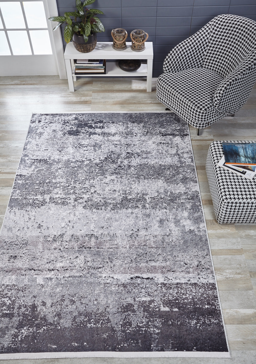 A modern living room featuring the Techno Flux - Washable Rug - JR1814 (Custom Size) in gray-anthracite, a black-and-white houndstooth armchair, matching ottoman with a book, and a white console table with plants against a blue wall. Ideal for busy spaces.