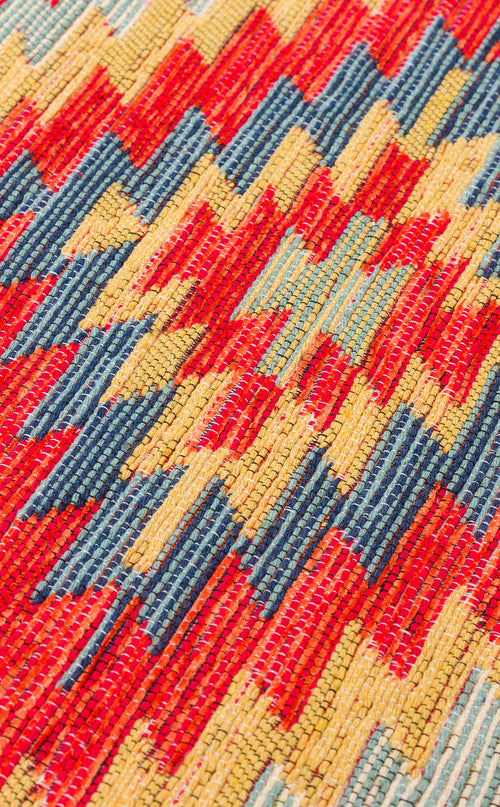 Load image 1 in gallery view. Close-up of Terra Thread - Kilim - NMD07, a flatwoven polyester fabric featuring a vibrant zigzag geometric pattern in red, yellow, and blue, inspired by tribal motifs. The detailed texture and weave are clearly visible.