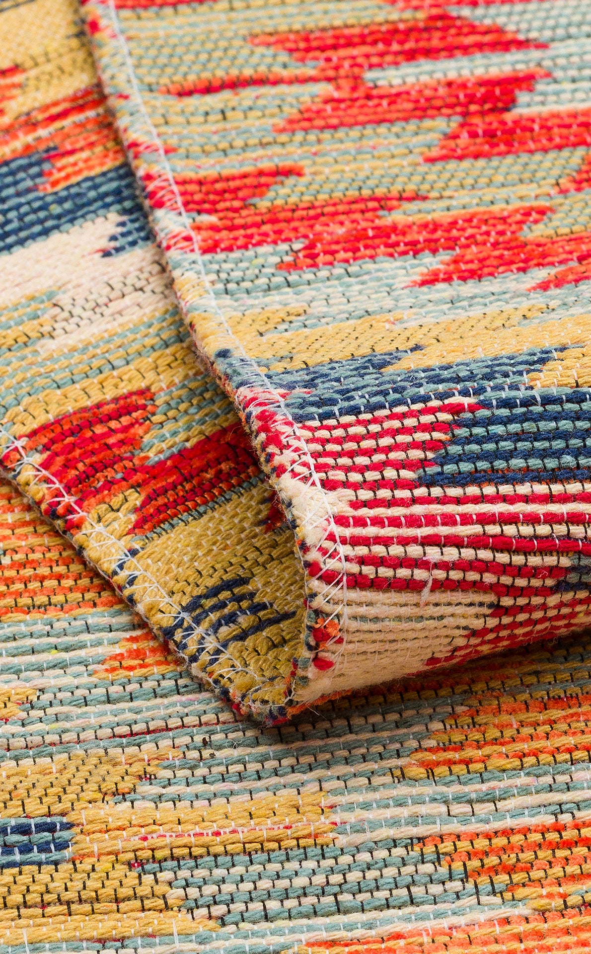 Close-up of the Terra Thread - Kilim - NMD07 rug featuring vibrant red, yellow, blue, and cream tribal patterns. A folded corner highlights its textured weave and detailed stitching along the edge.